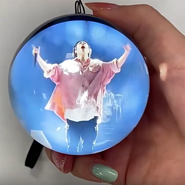 A hand holding a round, black-framed electronic music player or LED light displaying a video of a singer on a blue stage. Chinese text is overlaid at the top and bottom.