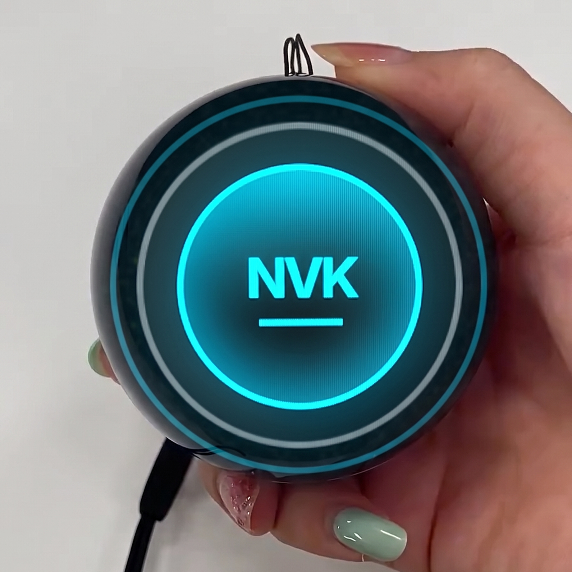 A hand holding a round black electronic device with a glowing blue circular logo that reads 'NVK' on the front, against a plain light background.